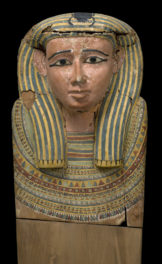 Carved wooden sculpture made of numerous pieces of wood joined with glue and doweled. Eyes and eyebrows may be glass inlays. This is the head and chest of a painted wooden coffin lid, showing the face of the deceased painted in ochre, wearing a long wig painted in blue and yellow stripes, and a large collar. On the top of the head a scarab beetle is painted.