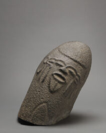 Ovoid volcanic stone monolith with face carved in low relief. Stylized face consists of a short nose, deeply set eyes, and an open mouth, the hair above shown closely and uniformly cropped. The sculpture measures 16-1/2 x 7 x 11 inches (41.9 x 17.8 x 27.9 cm).