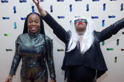 Two women in costumes have fun and strike poses in front of an IgNITE backdrop.