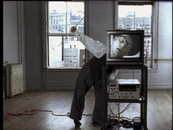 A video still of a person leaning over behind a television. The television depicts a man's face filling the screen that perfectly lines up with the person behind it.