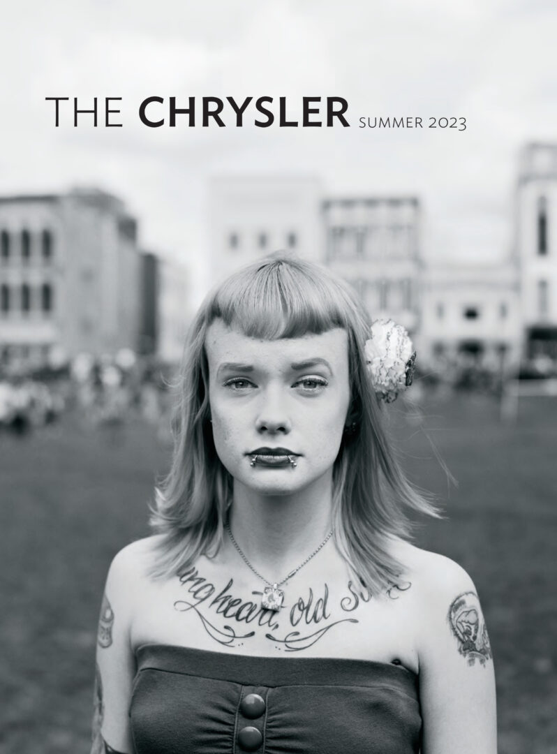 A photograph from the exhibition "Reckonings and Reconstructions: Southern Photography from the Do Good Fund" featuring a young woman with blunt bangs, lip piercings, and multiple piercings who stares transparently and vulnerably back at the viewer. Her chest tattoo reads "young heart, old soul."