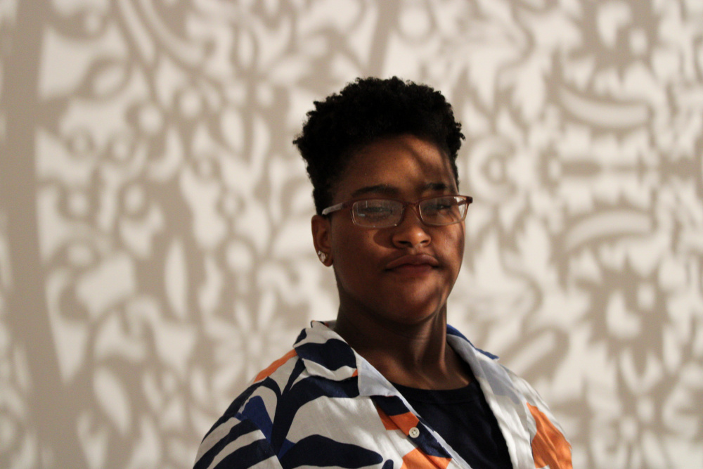2023 Summer Intern, K Barnes, stands in a gallery with the installation "All the Flowers Are for Me" by Anila Quayyum Agha. Shadow and light reflect on K's face.