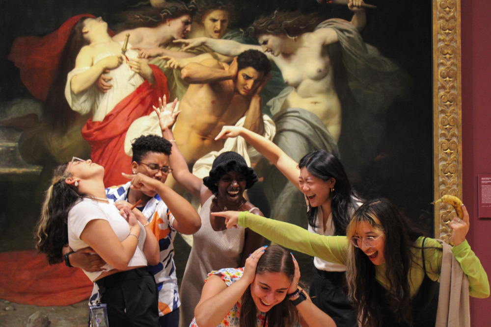 The 2023 Summer Interns act out the painting "Orestes Pursued by the Furies" by Adolphe-William Bouguereau. The painting is of Orestes,holding his hands over his ears with an expression of pain on his face, is swarthy and surrounded by four females with snakes for hair. The interns playfully stand in front of the dramatic painting and try to keep from laughing as they recreate the scene.