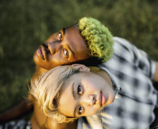 Photograph of a downward view of two young adults looking up at the camera. Although from different races and backgrounds, there is a sense of kinship evoked by the positioning of their heads and markers of a communal identity, such as hair, clothing etc.