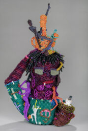 A teapot created out of glass beads featuring a central figure wearing glasses and smoking a cigarette surrounded by other beaded figures.